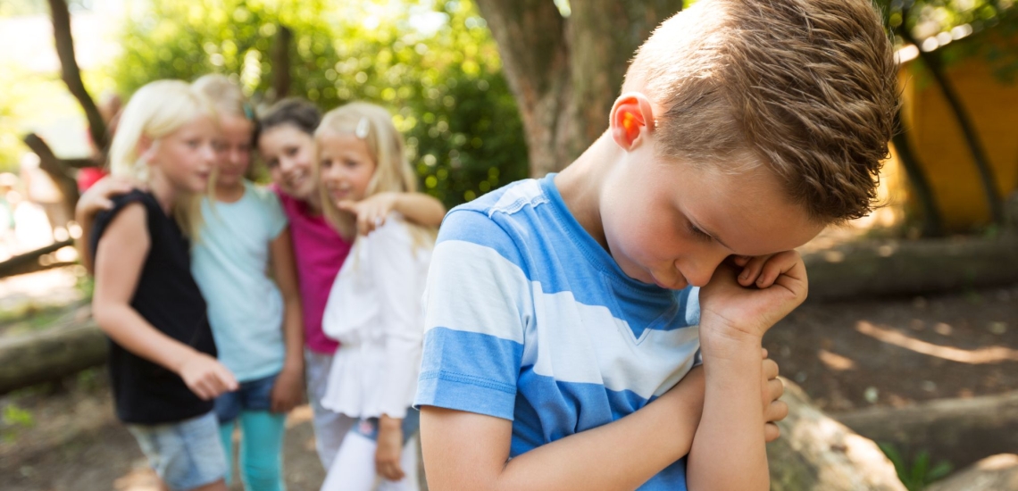 Junge traurig auf dem Schulhof, tuschelnde Gruppe Mädchen im Hintergrund