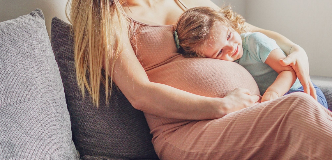 Schwangere Frau auf Sofa, Kleinkind legt Kopf auf ihren Bauch 