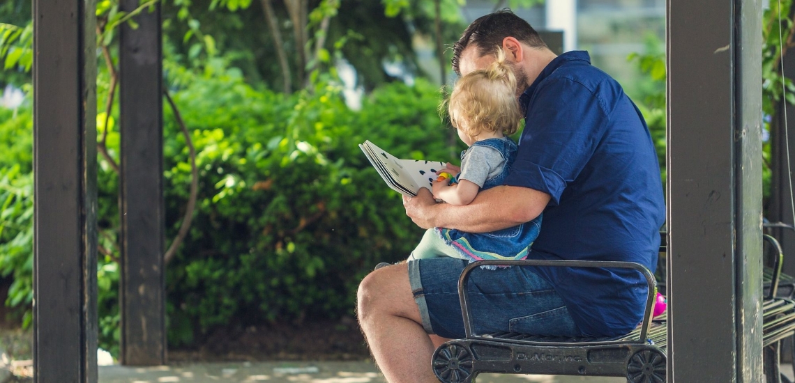 Vater sitzt mit Kind auf Bank und liest ihm vor 