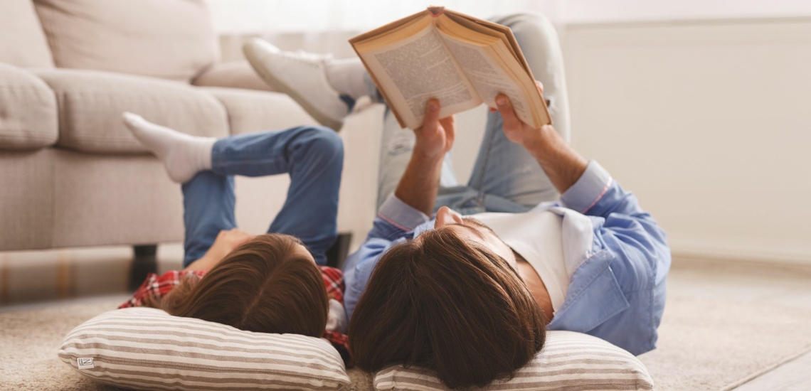 Vater und Kind liegen auf dem Boden auf Kissen und lesen gemeinsam in einem Buch 
