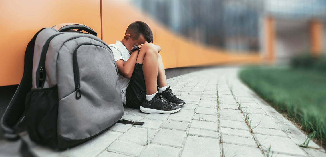 Junge mit Rucksack sitzt mit vor das Gesicht geschlagenen Händen auf dem Boden vor dem Schulgebäude 