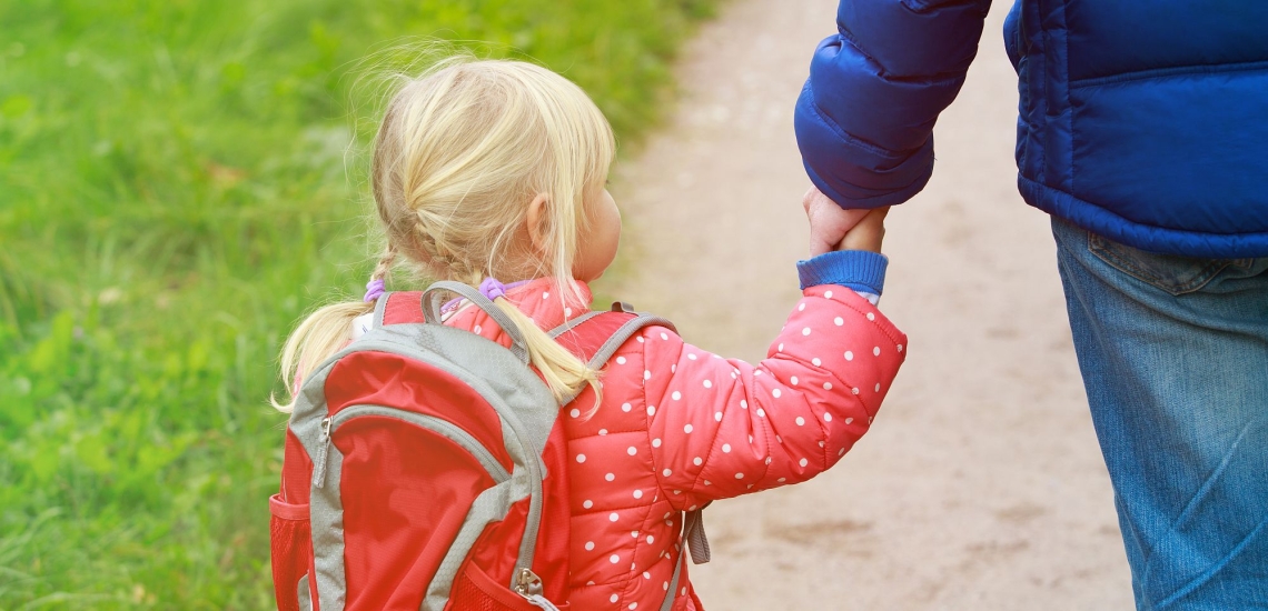 Kindergartenkind mit Rucksack geht an Hand des Vaters einen Weg entlang 