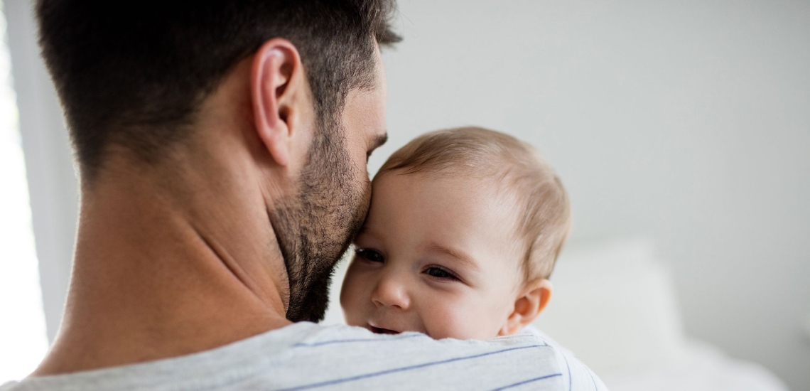 Vater hält kleines Kind auf dem Arm, das zufrieden lächelnd über seine Schulter schaut 