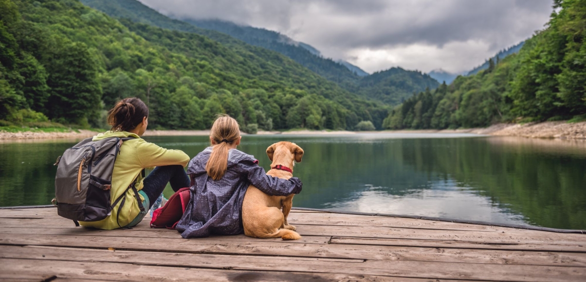 Frau, Kind und Hund sitzen auf Holzplanken und schauen auf einen Bergsee 