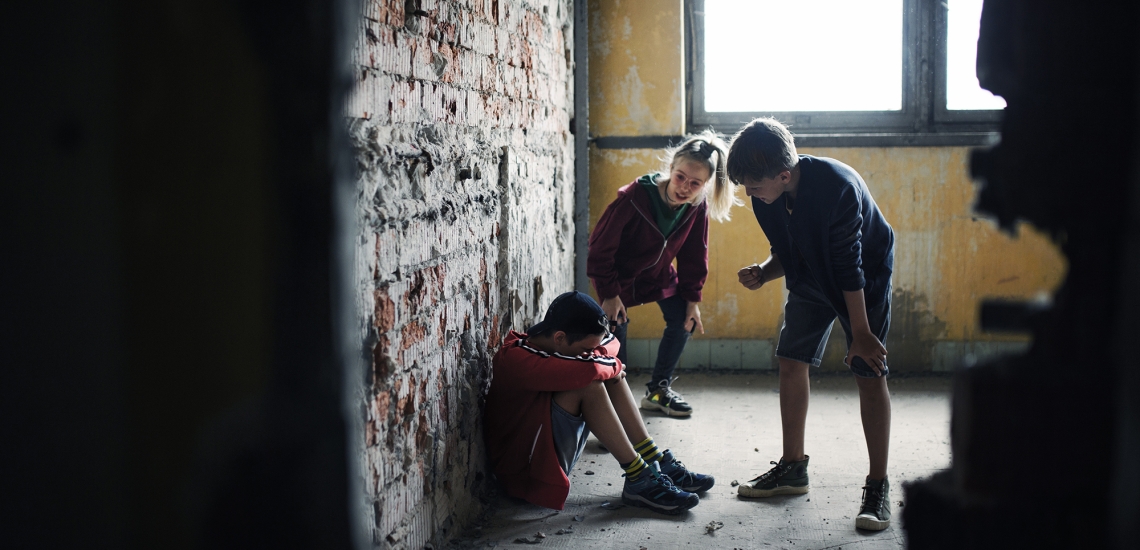 Ein Junge sitzt zusammengekauert auf dem Boden und hält schützend seine Arme vors Gesicht. Ein Mädchen und ein Junge stehen bedrohlich vor ihm.