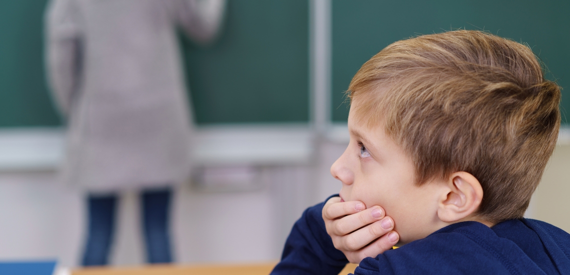 Eine Lehrerin schreibt etwas an die Tafel, ein Junge schaut nachdenklich zur Seite.