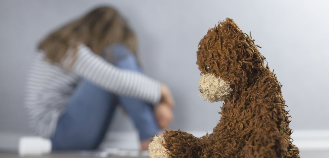 Ein Teddy sitzt im Vordergrund mit hängendem Kopf, hinter ihm - verschwommen zu sehen - sitzt ein in sich zusammengekauertes trauriges Mädchen.