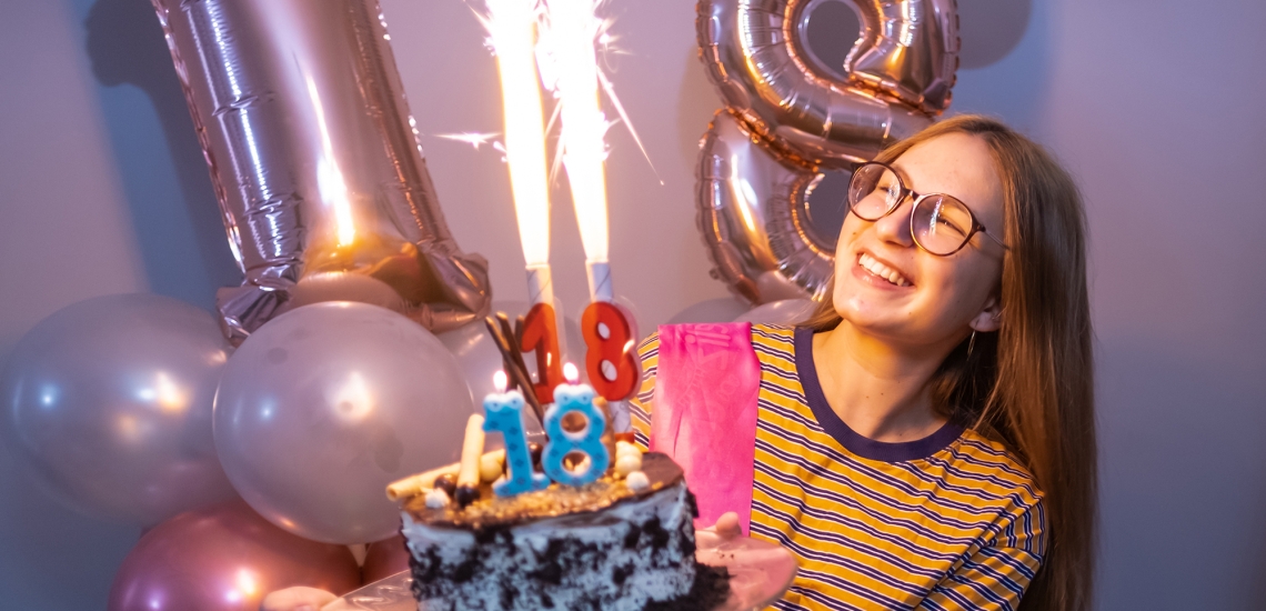 Ein Mädchen hält eine Torte in der Hand. Auf der Torte sind Geburtstagskerzen in Form einer 18, sprühendes Feuerwerk – und im Hintergrund sind Luftballons als Zahl 18 zu sehen.