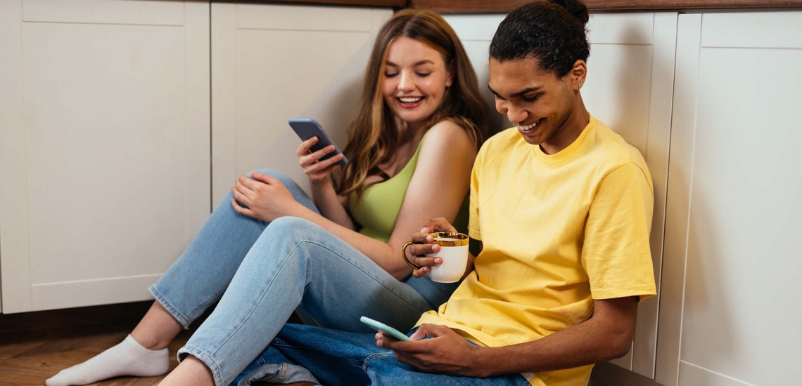 Ein junges Mädchen und ein junger Mann sitzen auf dem Küchenfußboden und schauen amüsiert auf ihre Smartphones.