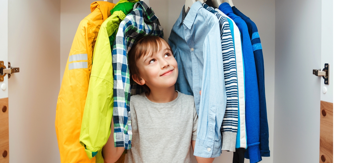 Ein Junge steht in seinem Kleiderschrank und schaut zwischen seinen Hemden auf der Kleiderstange hindurch.
