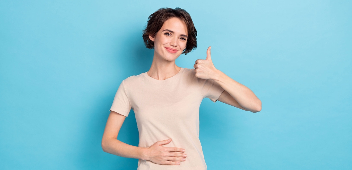 Eine junge Frau hält eine Hand auf ihren Bauch. Mit der anderen Hand macht sie ein Daumen-Hoch-Zeichen und lacht dabei.