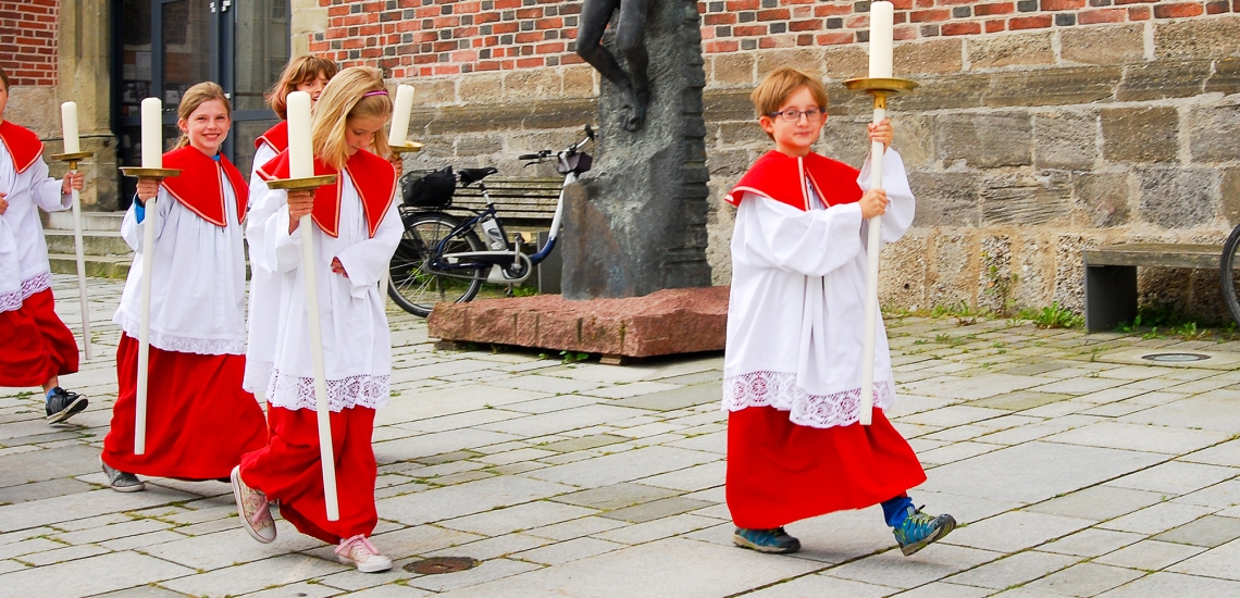 Junge Ministrantinnen und Ministranten laufen vor der Kirche dem Eingang entgegen und halten dabei Leuchten in der Hand.