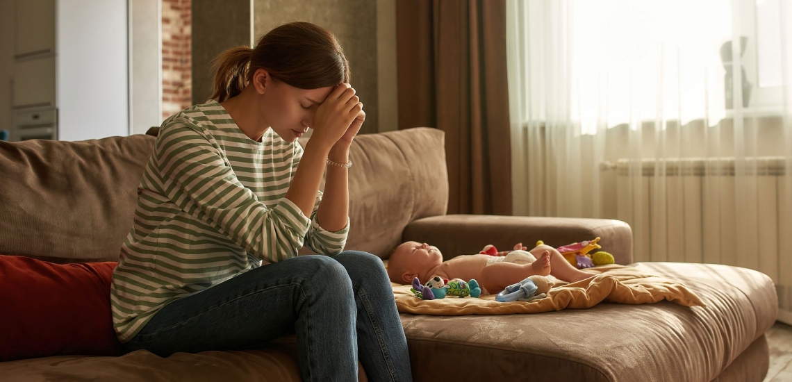 Mutter auf dem Sofa neben Säugling stützt bedrückt den Kopf auf ihre Hände 