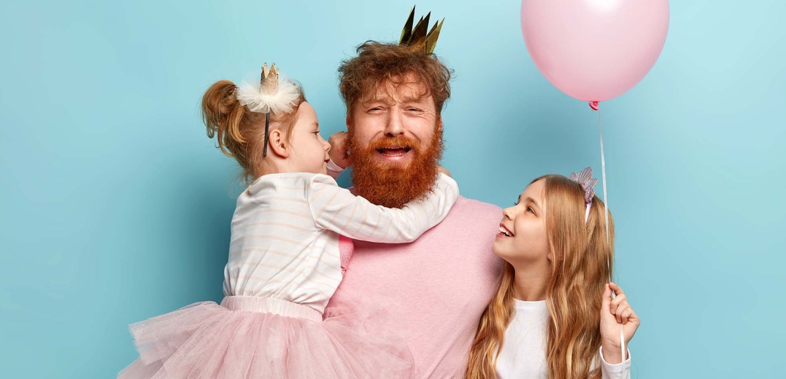 Ein Papa im rosa T-Shirt und mit goldener Krone hält seine eine Tochter im rosa Tütü auf dem Armm die andere Tochter steht mit einem rosa Luftballon neben ihm. Der Papa macht ein verzweifeltes Gesicht.