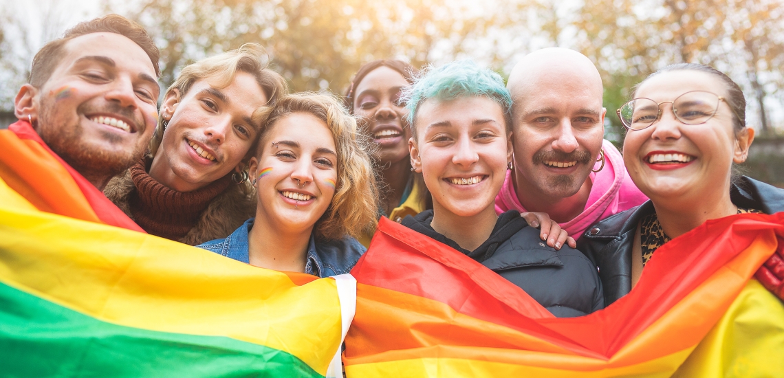 Queere Menschen halten eine Regenbogenfahne in der Hand und lächeln.