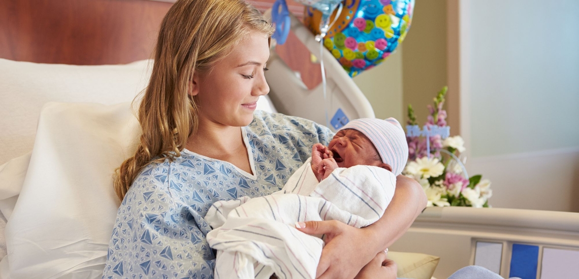 Teenagermutter mit neugeborenem Baby im Krankenhausbett mit Luftballons und Blumen 