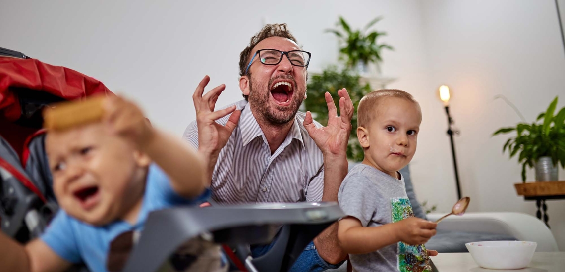 Zwei kleine Kinder essen, der Papa sitzt dazwischen, hebt seinen Kopf und schreit verzweifelt.