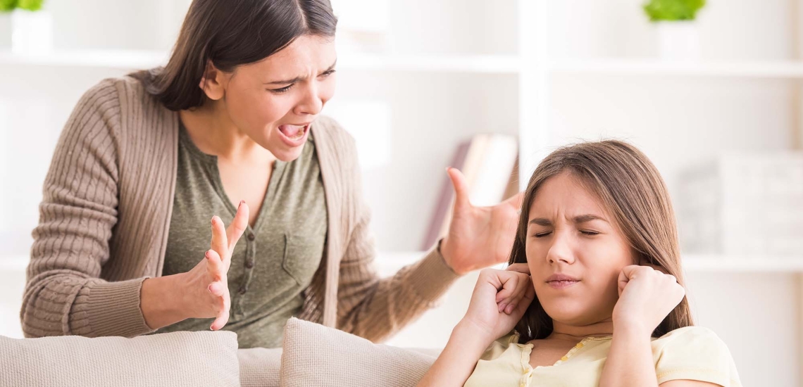 Eine Mutter schreit ihre Teenager-Tochter an, die auf dem Sofa sitzt und sich die Ohren zuhält.