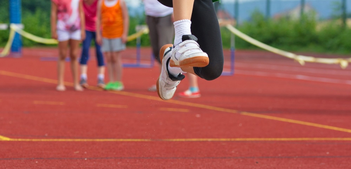 Kinderfüße in der Luft beim Weitsprung auf dem Sportplaz, im Hintergrund wartende Kinder