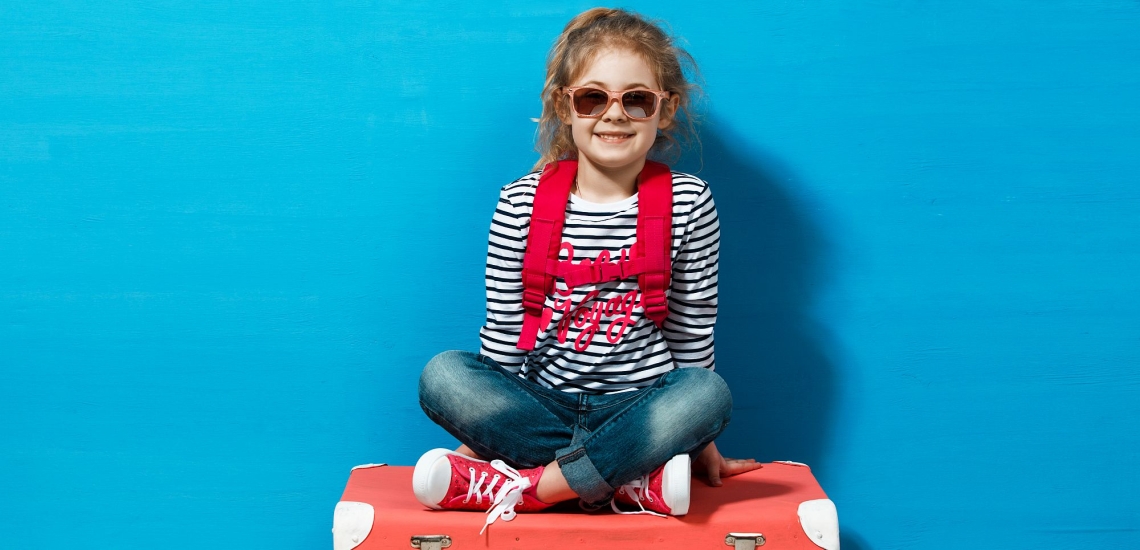 Mädchen mit Sonnenbrille und Rucksack sitzt lachend auf Koffer 