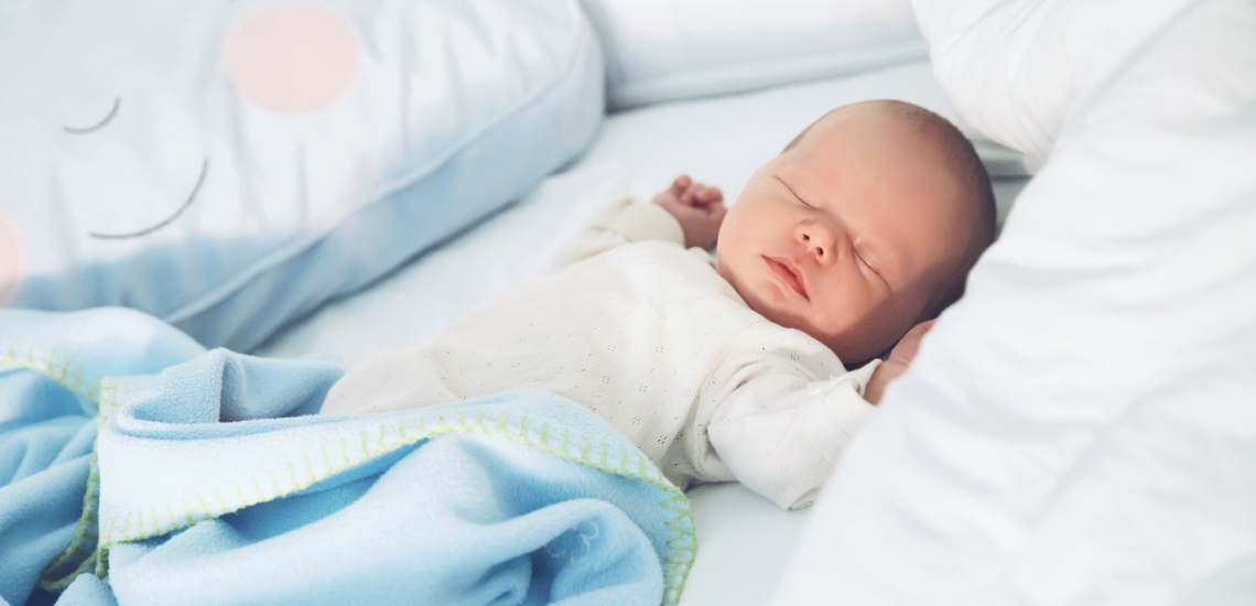 Säugling im Strampelanzug liegt zugedeckt auf Bett zwischen Kissen 