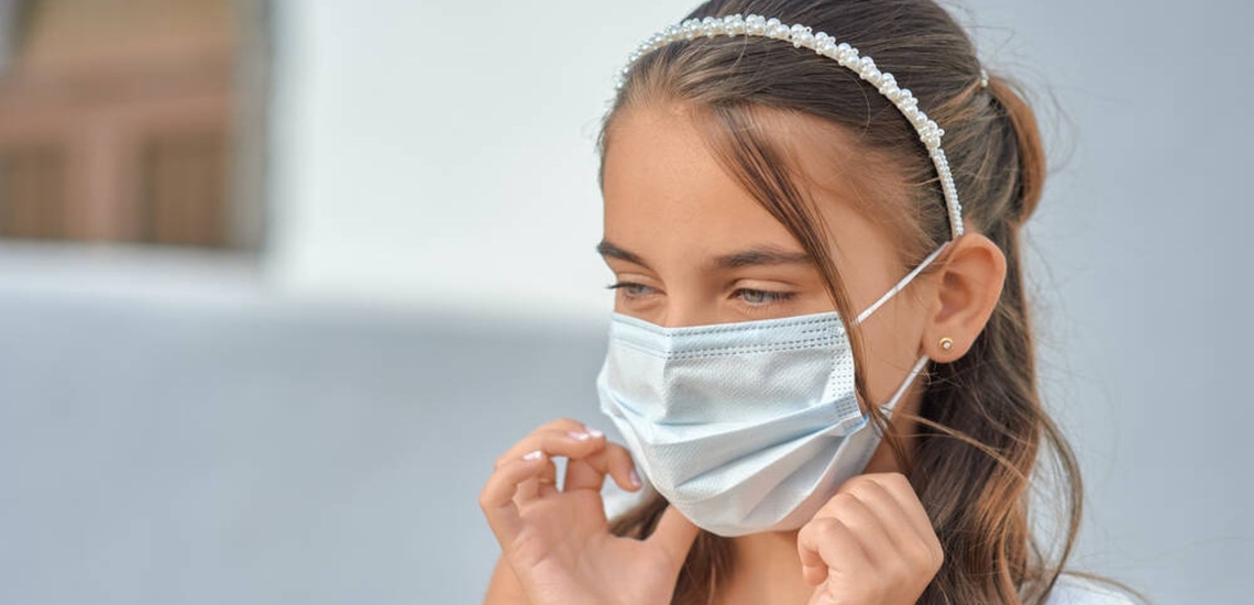 Erstkommunionkind in Coronapandemie im weißen Kleid mit Maske 