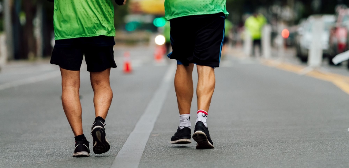 Zwei Männer von hinten joggen über Straße mit Laufschuhen und grünen Warnwesten 