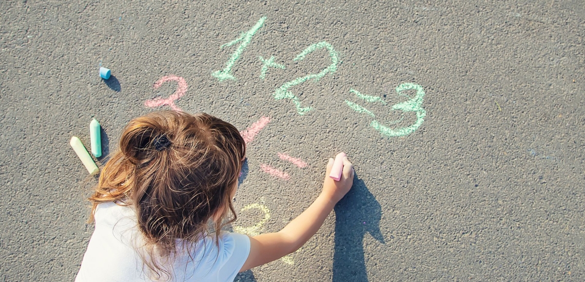 Mädchen schreibt mit Kreide Rechenaufgaben auf Straße 