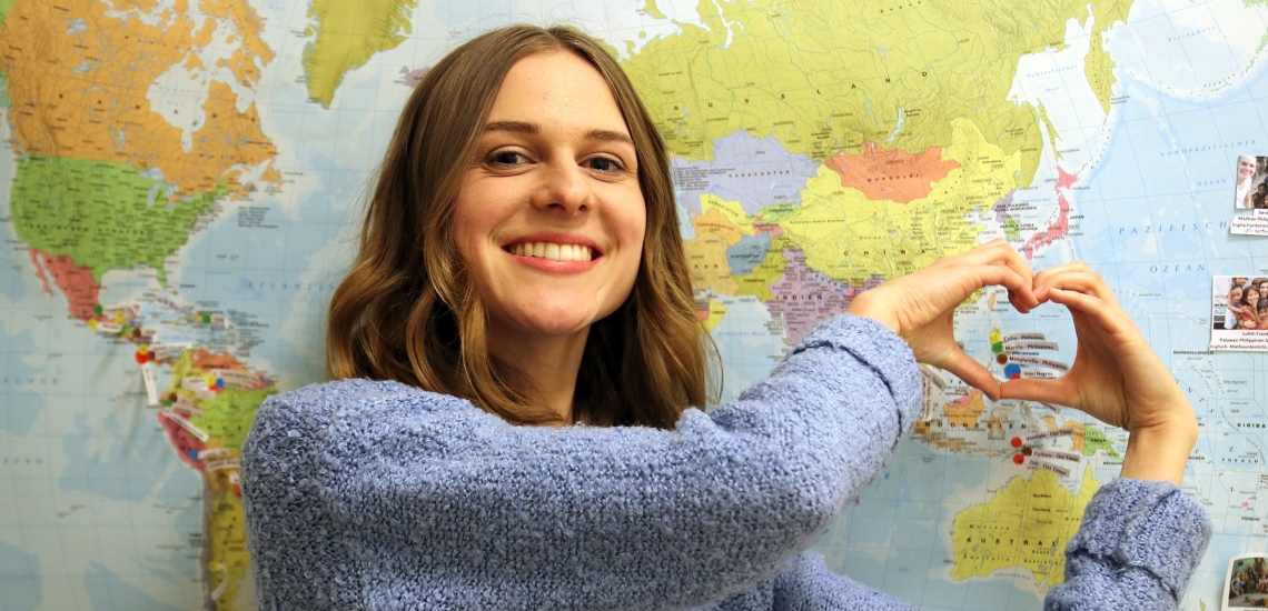 Sara Straub formt vor Weltkarte mit den Händen ein Herz 