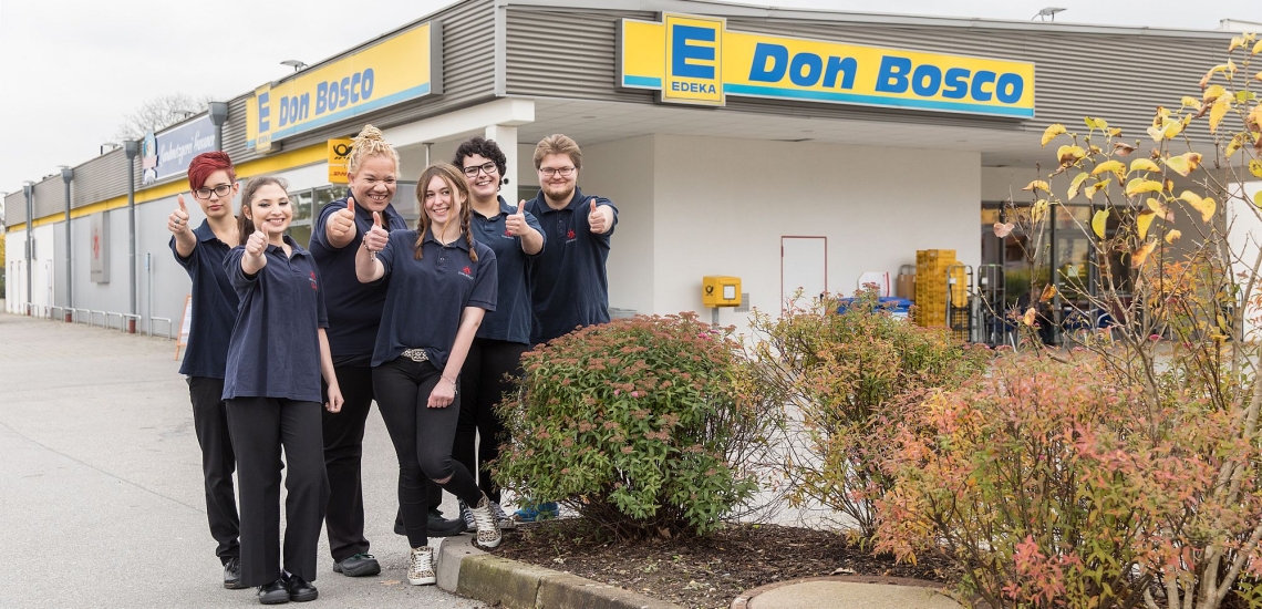 Sechs Jugendliche in dunkelblauen Don Bosco Shirts zeigen Daumen hoch vor Don Bosco Edeka Markt 