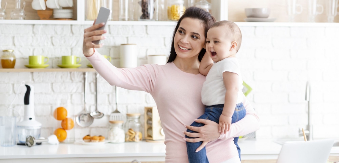 Mutter macht Selfie von sich mit Baby auf dem Arm 