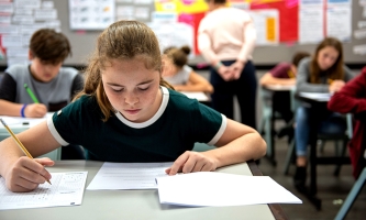 Eine Jugendliche mit grünem Shirt sitzt an einem Tisch im Klassenraum einer Schule und schreibt einen Test. Im Hintergrund sind Mitschülerinnen und Mitschüler in der gleichen Situation zu sehen.