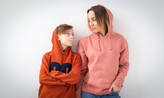 Ein männlicher Teenager und seine Mutter stehen in Kapuzenpullovern vor einer weißen Wand und blicken sich ind ie Augen. Der Teenager hat die Arme verschränkt.