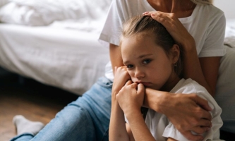 Trauriges Kind wird von Mutter im Arm gehalten, beide sitzen auf Boden vor Bett