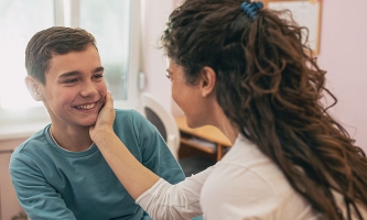 Mutter knuddelt Sohn vergnügt an der Wange, Sohn lächelt sie an.