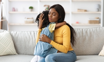Eine Mutter umarmt ihre Tochter. Sie trägt ein gelbes Longsleeve. Die Tochter ist ca. 5 Jahre alt und trägt ein blaues Kleid über einem gelben T-Shirt.
