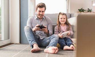 Ein Vater spielt mit seiner Tochter ein Computerspiel, beide halten eine Spielkonsole in der Hand.