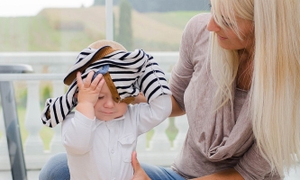 Kleinkind zieht sich alleine den Pullover über den Kopf, Mutter hält Kind und schaut zu