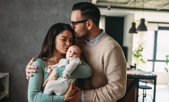 Eine Frau hält ein sehr junges Baby im Arm und küsste es auf die Stirn. Ein Mann hält umarmt die Frau und legt seine Hände dabei auf ihre Schulter und ihre Hand, die das Baby hält. Dabei küsst er sie auf den Kopf.
