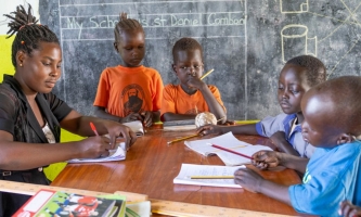Frau mit Kindern am Tisch mit Heften und Büchern, im Hintergrund eine Tafel