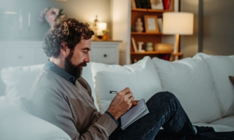 Mann mit Bart sitzt nachdenklich auf Sofa in gemütlichem Wohnzimmer mit Stift und Notizblock