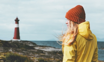 Eine junge Frau steht mit gelber Reckenjacke und oranger Mütze am Meer, im Hintergrund ist ein Leuchtturm zu sehen.