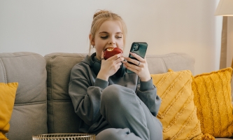Ein blondes Mädchen sitzt apfelessend auf der Couch und schaut aufs Handy.