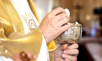 Priester hat Kelch mit Hostien in einer Hand und zeigt mit der anderen eine Hostie