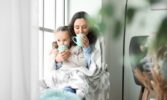 Eine Mutter hat ihre Tochter auf dem Schoß. Beide sind in eine Decke eingekuschelt und trinken heißen Kakao.