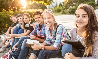 Eine gemischte Gruppe aus Schülerinnen und Schülern sitzt bei tief stehender Sonne auf Stufen im Freien. Sie haben Bücher, Notizhefte und ein Tablet in der Hand und lächeln in die Kamera.