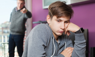 Vater steht im Hintergrund und Zeit aufgebracht mit dem Finger auf den jugendlichen Jungen, der ihm den Rücken zudreht.