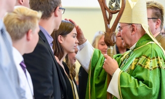 Ein Bischof zeichnet mit Chrisamöl ein Kreuz auf die Stirn einer Jugendlichen, die in einer Reihe mit anderen Firmlingen steht