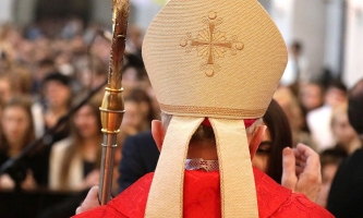 Bischof mit Mitra und Bischofsstab steht vor einer Jugendlichen und hebt die Hand, um ihr ein Kreuz auf die Stirn zu zeichnen