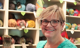Frau mit kurzen blonden Haaren und Brille vor Regalen mit Wollknäueln